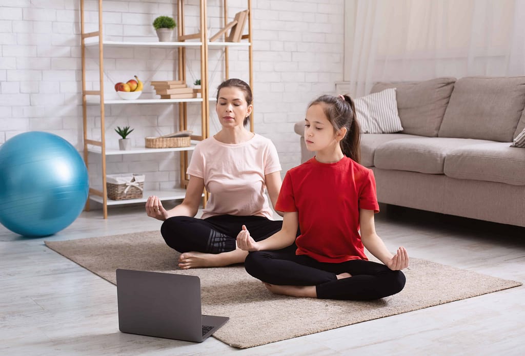 Home yoga workout. Mom and teen daughter meditating to online tutorial on laptop in living room